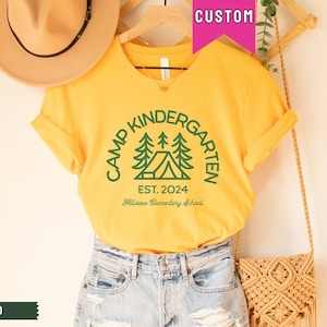 May include: A yellow t-shirt with a green graphic of a tent and trees. The text "Camp Kindergarten Est. 2024 Hillview Elementary School" is printed on the shirt.