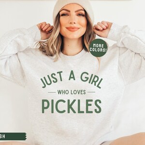 May include: A light grey sweatshirt with the text "Just a girl who loves pickles" in green. The text is in a circular design with a green border. The sweatshirt has a "More Colors!" tag in a green circle.