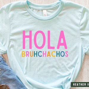 May include: A light blue t-shirt with the text "HOLA BRUHCHACHOS" in pink, yellow, green, blue, and orange letters.