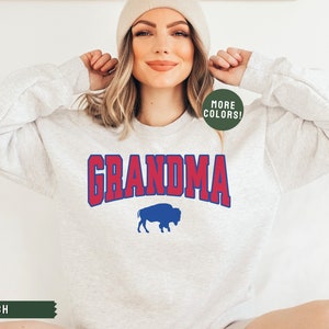 May include: A light grey sweatshirt with the word "GRANDMA" in red and blue lettering with a blue buffalo silhouette underneath. The sweatshirt is being modeled by a person wearing a beige beanie.