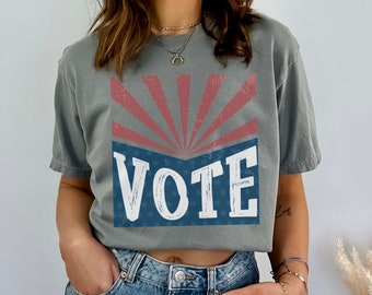 May include: A grey t-shirt with a red, white, and blue graphic that says "VOTE" with a sunburst design behind the text.