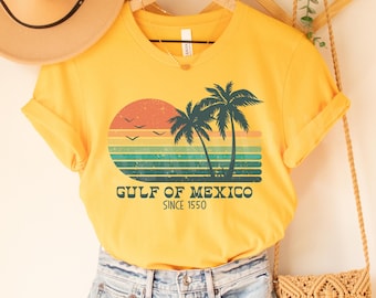 Pode incluir: Camiseta amarela de manga curta com um gráfico de pôr do sol vintage e palmeiras. O texto "GULF OF MEXICO SINCE 1550" está impresso na camiseta.