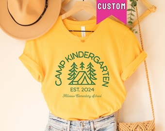 May include: A yellow t-shirt with a green graphic of a tent and trees. The text "Camp Kindergarten Est. 2024 Hillview Elementary School" is printed on the shirt.