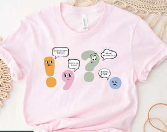 May include: A soft pink t-shirt with a graphic design featuring punctuation marks with faces and speech bubbles. The text in the speech bubbles reads "Seriously, Bruh!" "Hold Up, Bruh" "Bruh, Actually?" and "Bruh, Stop."