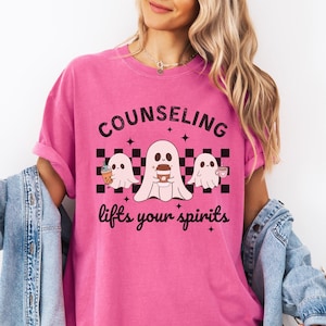 May include: Pink t-shirt with a black and white graphic of three ghosts holding coffee cups. The text "Counseling lifts your spirits" is printed on the shirt.
