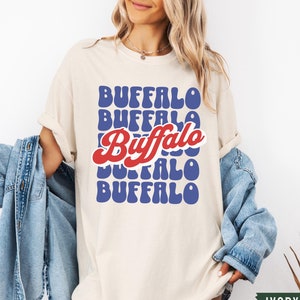 May include: A cream colored t-shirt with a retro style graphic that says "Buffalo" in blue and red letters.