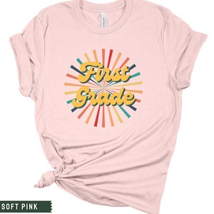 May include: A soft pink t-shirt with a retro-style graphic that says "First Grade" in yellow letters with a colorful sunburst design.