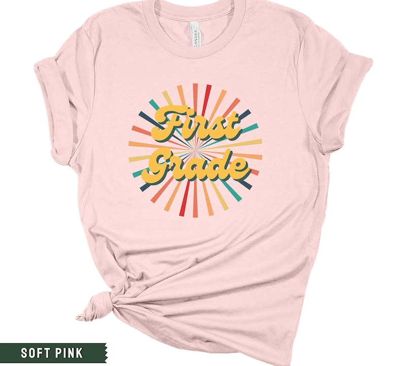 May include: A soft pink t-shirt with a retro-style graphic that says "First Grade" in yellow letters with a colorful sunburst design.