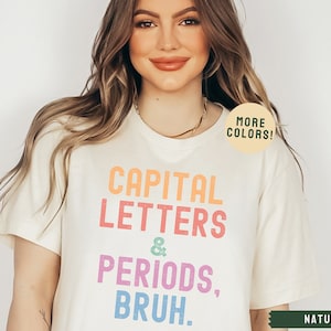 Puede incluir: Una camiseta blanca con el texto "CAPITAL LETTERS & PERIODS, BRUH." en colores arco&iacute;ris. El texto est&aacute; en negrita, fuente sin serifas. La camiseta est&aacute; siendo modelada por una persona con cabello largo y casta&ntilde;o. El texto "MORE COLORS!" est&aacute; en un c&iacute;rculo verde en el hombro derecho de la camiseta. El texto "NATURAL" est&aacute; en verde en la esquina inferior derecha de la imagen.