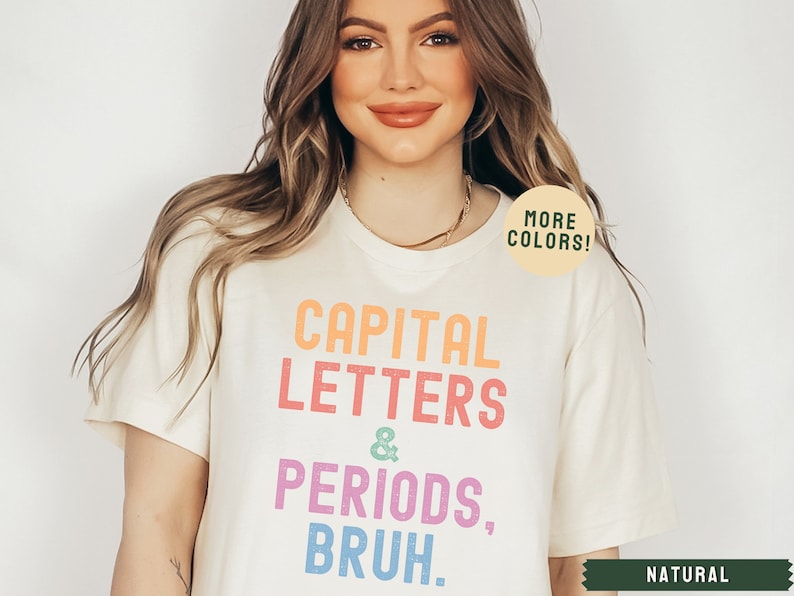 Puede incluir: Una camiseta blanca con el texto "CAPITAL LETTERS & PERIODS, BRUH." en colores arco&iacute;ris. El texto est&aacute; en negrita, fuente sin serifas. La camiseta est&aacute; siendo modelada por una persona con cabello largo y casta&ntilde;o. El texto "MORE COLORS!" est&aacute; en un c&iacute;rculo verde en el hombro derecho de la camiseta. El texto "NATURAL" est&aacute; en verde en la esquina inferior derecha de la imagen.