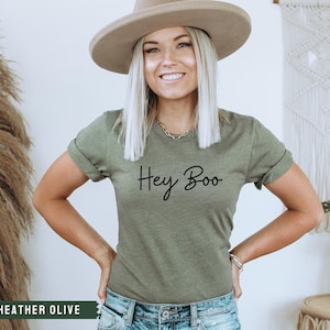 May include: A woman wearing a heather olive t-shirt with the text "Hey Boo" printed on the front. She is wearing a wide-brimmed hat and jeans.
