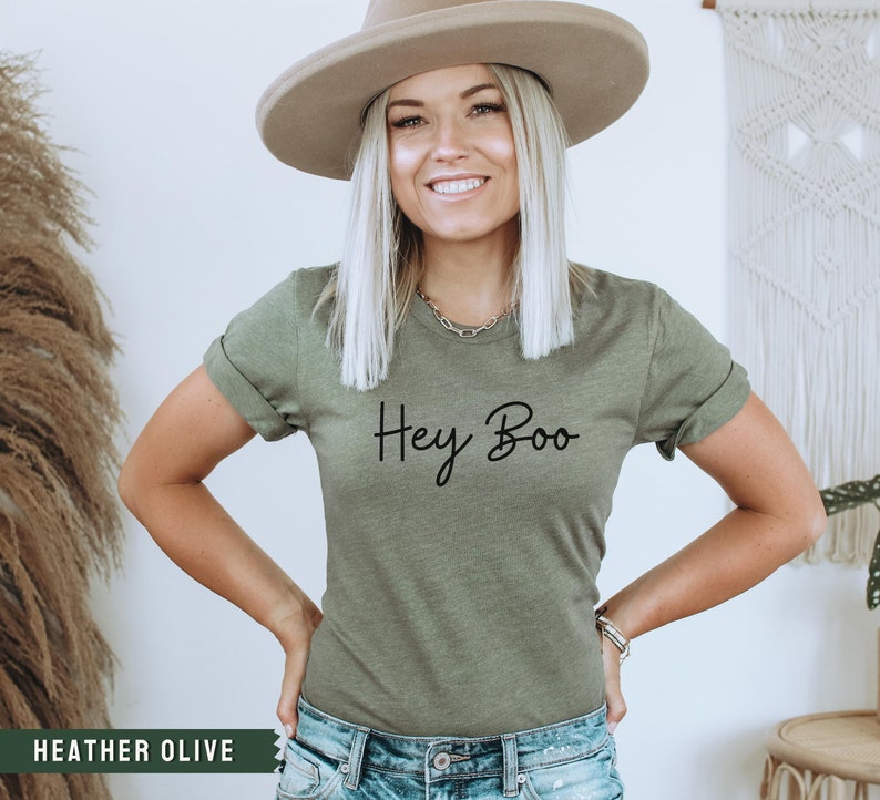 May include: A woman wearing a heather olive t-shirt with the text "Hey Boo" printed on the front. She is wearing a wide-brimmed hat and jeans.