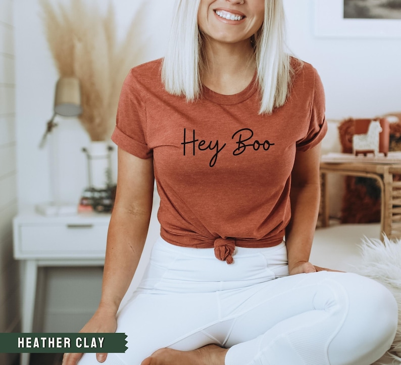 May include: A woman wearing a rust-colored t-shirt with the text "Hey Boo" in black script. She is wearing white pants and is sitting with her legs crossed.