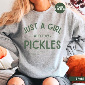 May include: A grey sweatshirt with the text "Just a girl who loves pickles" in green lettering. The sweatshirt is being worn by a person. The text "More Colors!" is in a green circle in the top right corner of the image. The text "Sport Grey" is in the bottom right corner of the image.