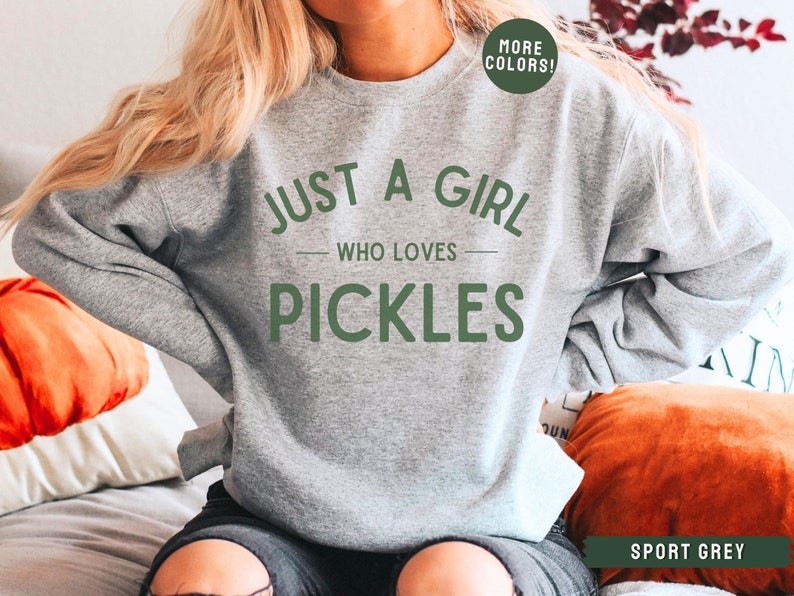 May include: A grey sweatshirt with the text "Just a girl who loves pickles" in green lettering. The sweatshirt is being worn by a person. The text "More Colors!" is in a green circle in the top right corner of the image. The text "Sport Grey" is in the bottom right corner of the image.