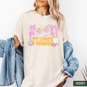 Peut inclure: Un t-shirt blanc avec le texte "Spooky Teacher" en lettres roses et orange. Le texte est entour&eacute; d'une boule disco, d'un fant&ocirc;me et d'autres graphismes sur le th&egrave;me d'Halloween.