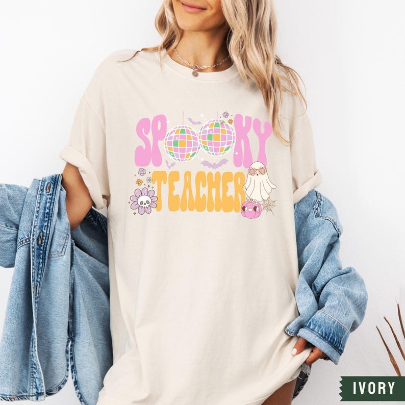 Peut inclure: Un t-shirt blanc avec le texte "Spooky Teacher" en lettres roses et orange. Le texte est entour&eacute; d'une boule disco, d'un fant&ocirc;me et d'autres graphismes sur le th&egrave;me d'Halloween.