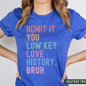 Puede incluir: Una camiseta azul real con el texto "ADMIT IT YOU LOW KEY LOVE HISTORY, BRUH" en colores arco&iacute;ris.