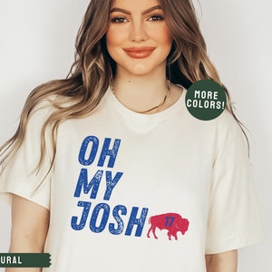 May include: A white t-shirt with blue text that reads "Oh My Josh" and a blue and red buffalo with the number 17 on it. The text has a distressed look. The shirt is on a model with long brown hair.