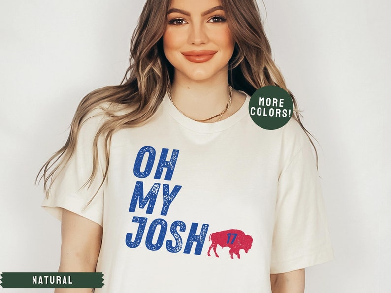 May include: A white t-shirt with blue text that reads "Oh My Josh" and a blue and red buffalo with the number 17 on it. The text has a distressed look. The shirt is on a model with long brown hair.
