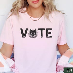 Puede incluir: Una camiseta rosa suave con la palabra "VOTE" impresa en negro con un gr&aacute;fico de gato negro en el medio de la palabra.