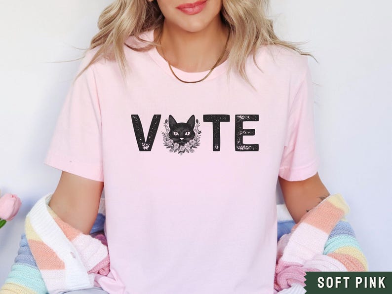 Puede incluir: Una camiseta rosa suave con la palabra "VOTE" impresa en negro con un gr&aacute;fico de gato negro en el medio de la palabra.