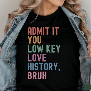 Puede incluir: Una camiseta negra con el texto "ADMIT IT YOU LOW KEY LOVE HISTORY, BRUH" en colores arco&iacute;ris. El texto est&aacute; en una fuente desgastada.
