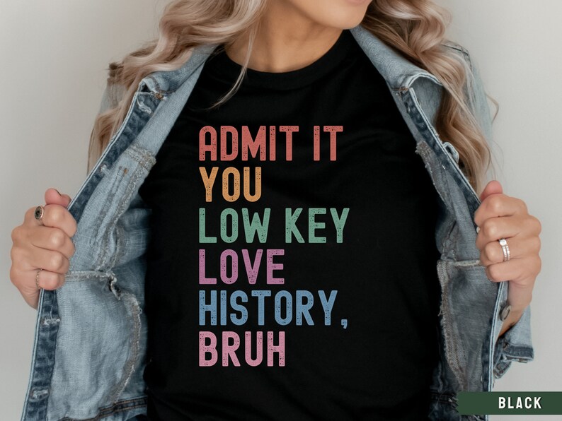 Puede incluir: Una camiseta negra con el texto "ADMIT IT YOU LOW KEY LOVE HISTORY, BRUH" en colores arco&iacute;ris. El texto est&aacute; en una fuente desgastada.