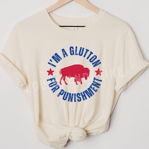 May include: Cream-colored t-shirt with a graphic design. The design features a red buffalo inside a blue circle with the text "I'M A GLUTTON FOR PUNISHMENT" in blue. Two red stars are on either side of the buffalo.