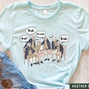 Peut inclure: T-shirt bleu clair avec un graphique des P&egrave;res fondateurs assis autour d'une table avec des bulles de dialogue qui disent "Bruh."