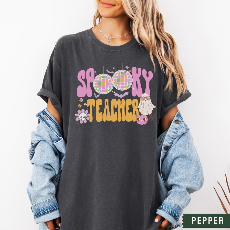 Peut inclure: Un t-shirt gris fonc&eacute; avec le texte "Spooky Teacher" dans une police r&eacute;tro. Le design comprend un fant&ocirc;me, des boules disco, des chauves-souris et des fleurs.