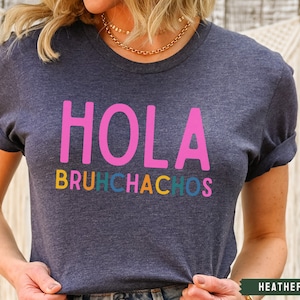 May include: A heather navy blue t-shirt with the text "Hola Bruchhachos" in pink, orange, yellow, green, and blue lettering.