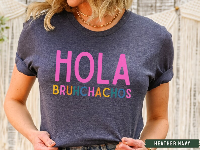 May include: A heather navy blue t-shirt with the text "Hola Bruchhachos" in pink, orange, yellow, green, and blue lettering.