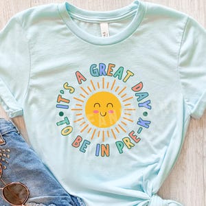 Peut inclure: Un t-shirt bleu clair &agrave; manches courtes avec un motif central repr&eacute;sentant un soleil jaune souriant entour&eacute; du texte "IT'S A GREAT DAY TO BE IN PRE-K" en lettres arrondies color&eacute;es. Le design utilise des tons de bleu, jaune, rose et orange.