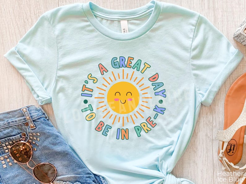 Peut inclure: Un t-shirt bleu clair &agrave; manches courtes avec un motif central repr&eacute;sentant un soleil jaune souriant entour&eacute; du texte "IT'S A GREAT DAY TO BE IN PRE-K" en lettres arrondies color&eacute;es. Le design utilise des tons de bleu, jaune, rose et orange.