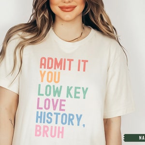 Puede incluir: Una camiseta blanca con el texto "ADMIT IT YOU LOW KEY LOVE HISTORY, BRUH" impreso en colores arco&iacute;ris. El texto est&aacute; en negrita, con una fuente de bloque. La camiseta est&aacute; en una modelo y la palabra "NATURAL" es visible en la esquina inferior derecha.