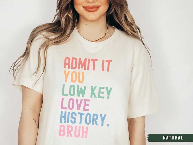 Puede incluir: Una camiseta blanca con el texto "ADMIT IT YOU LOW KEY LOVE HISTORY, BRUH" impreso en colores arco&iacute;ris. El texto est&aacute; en negrita, con una fuente de bloque. La camiseta est&aacute; en una modelo y la palabra "NATURAL" es visible en la esquina inferior derecha.