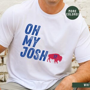 May include: White t-shirt with blue text that reads "Oh My Josh" and a red buffalo with the number 17 on it. The text has a distressed look.  The shirt is available in other colors.