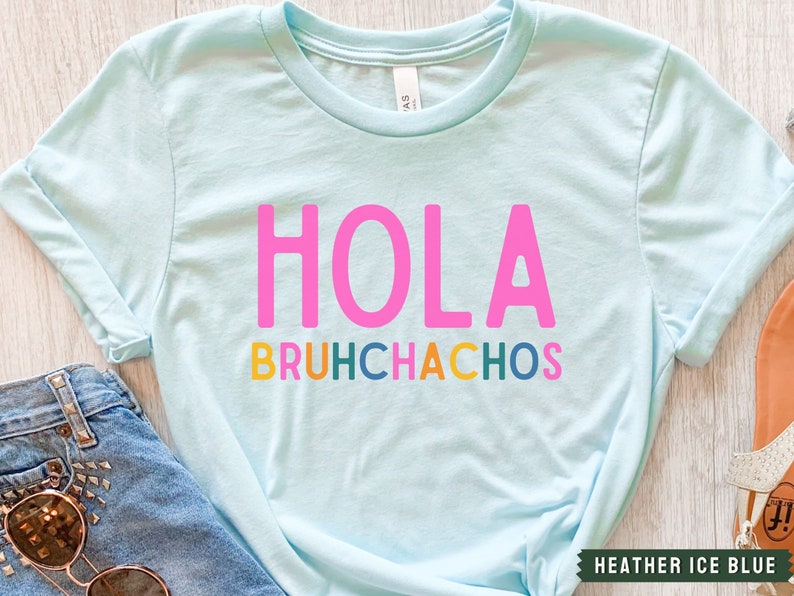 May include: A light blue t-shirt with the text "HOLA BRUHCHACHOS" in pink, yellow, green, blue, and orange letters.