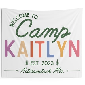 May include: A white tapestry with green text that reads "Welcome to Camp Kaitlyn" in a colorful rainbow font. The text "EST. 2023" and "Adirondack Mts." are also included in green text. The tapestry measures 60 inches by 50 inches.
