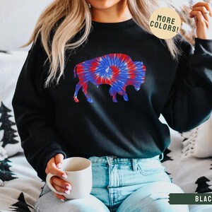 May include: Black sweatshirt featuring a tie-dye buffalo design in red, white, and blue. The sweatshirt has a crew neck and long sleeves. The text "MORE COLORS!" is in a circle on the top right. The text "BLACK" is in a banner at the bottom.