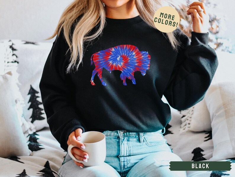 May include: Black sweatshirt featuring a tie-dye buffalo design in red, white, and blue. The sweatshirt has a crew neck and long sleeves. The text "MORE COLORS!" is in a circle on the top right. The text "BLACK" is in a banner at the bottom.