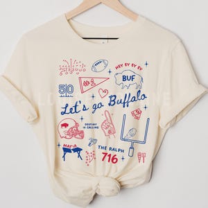 May include: Cream-colored t-shirt with a graphic design featuring the text "Let's go Buffalo" and various football-related illustrations in red and blue. Includes a football, helmet, and the number 716.