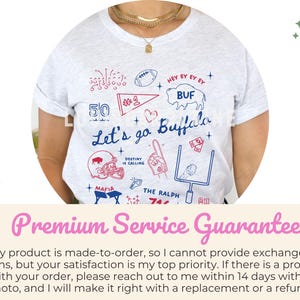May include: A light gray t-shirt with a hand-drawn design featuring the text "Let's go Buffalo" in blue script. The design includes a football, a helmet, and other football-related graphics in red and blue.