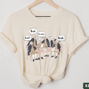 Peut inclure: Un t-shirt cr&egrave;me avec un graphique repr&eacute;sentant les P&egrave;res fondateurs assis autour d'une table avec des bulles de dialogue au-dessus de leurs t&ecirc;tes qui disent "Bruh." et "Bruh?" Le graphique est dans un style cartoon.
