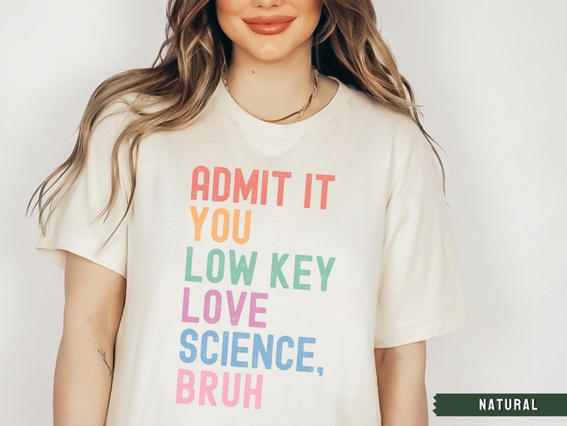 May include: A white t-shirt with the text "ADMIT IT YOU LOW KEY LOVE SCIENCE, BRUH" printed in rainbow colors. The text is in a vertical stack. The shirt is on a model and the color of the shirt is "NATURAL".