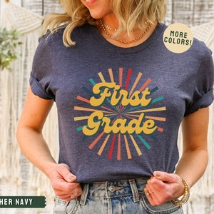 May include: A heather navy blue t-shirt with a retro-style sunburst design and the text "First Grade" in yellow. The text is in a bold, cursive font. The sunburst is made up of colorful rays in shades of red, orange, yellow, green, and blue.