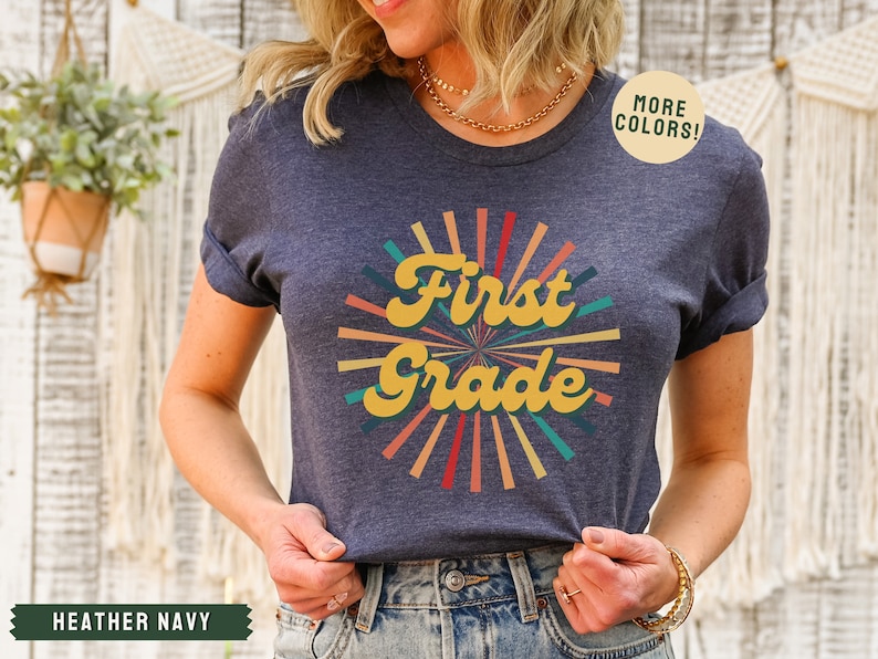 May include: A heather navy blue t-shirt with a retro-style sunburst design and the text "First Grade" in yellow. The text is in a bold, cursive font. The sunburst is made up of colorful rays in shades of red, orange, yellow, green, and blue.