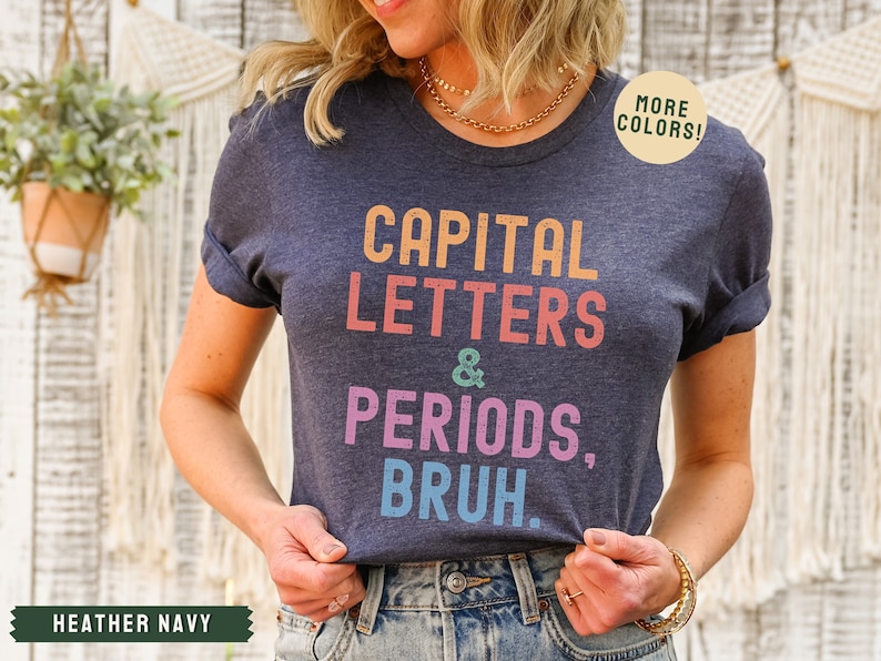 Puede incluir: Una camiseta azul marino jaspeada con el texto "CAPITAL LETTERS & PERIODS, BRUH." en colores arco&iacute;ris. El texto est&aacute; en un estilo de fuente retro.