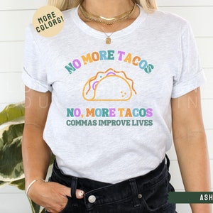 May include: A white t-shirt with the text "NO MORE TACOS NO, MORE TACOS COMMAS IMPROVE LIVES" in colourful letters. The shirt features a graphic of a taco in the centre.